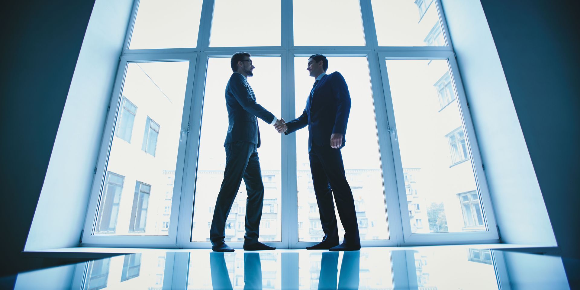 Two businessmen shaking hands in front of a large window, possibly after a deal.
