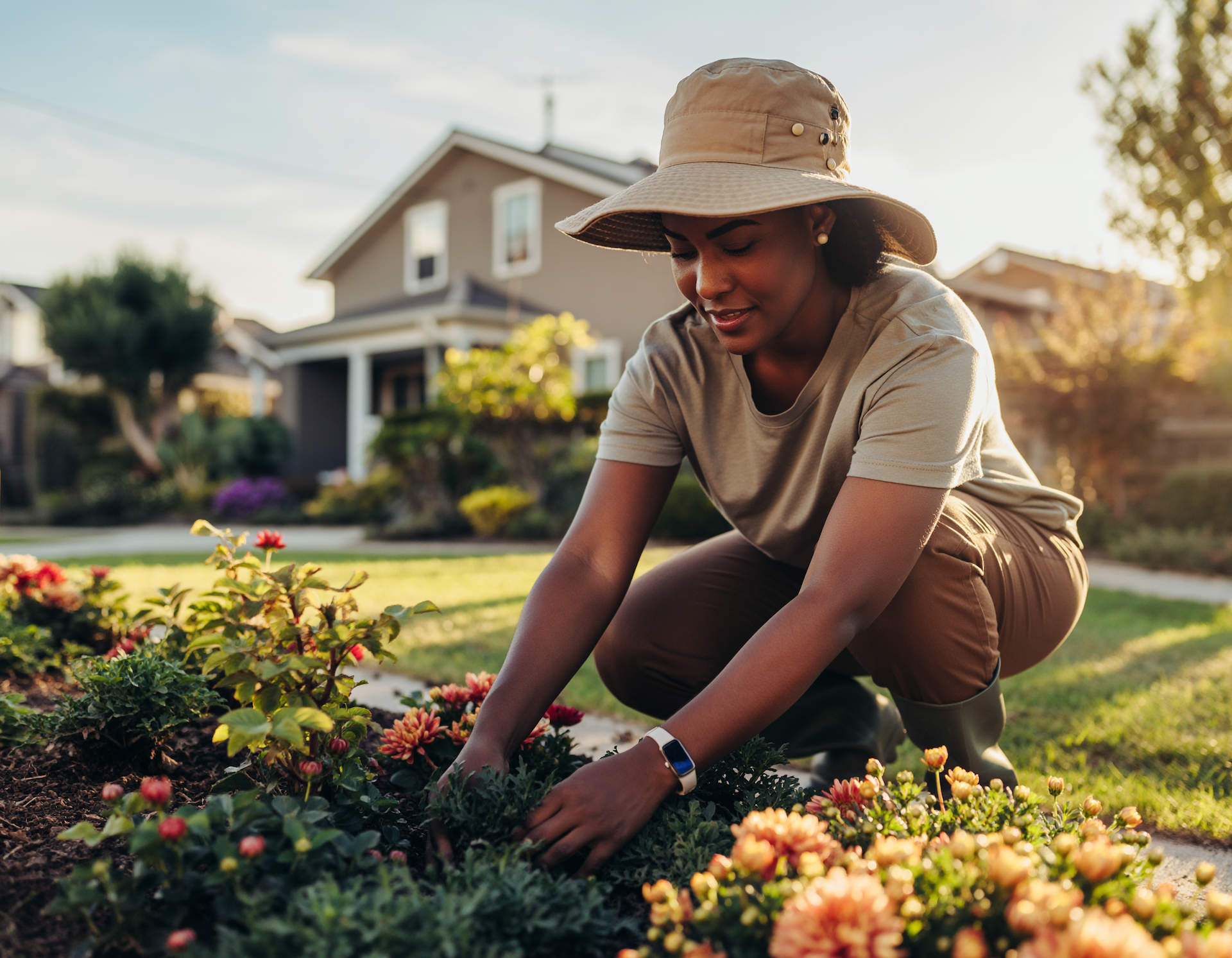 Woman in a hat tending to flowers in a garden bed in front of a house.