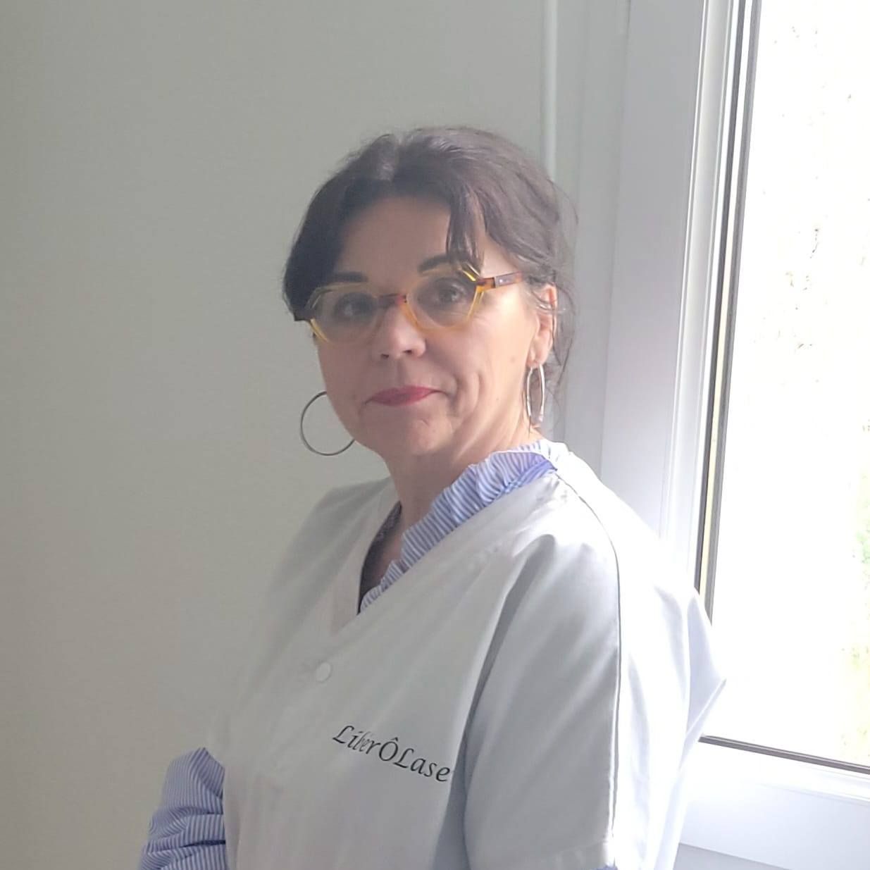 Femme en blouse blanche, lunettes et créoles, debout près d'une fenêtre. Il s'agit probablement d'une professionnelle de la santé.