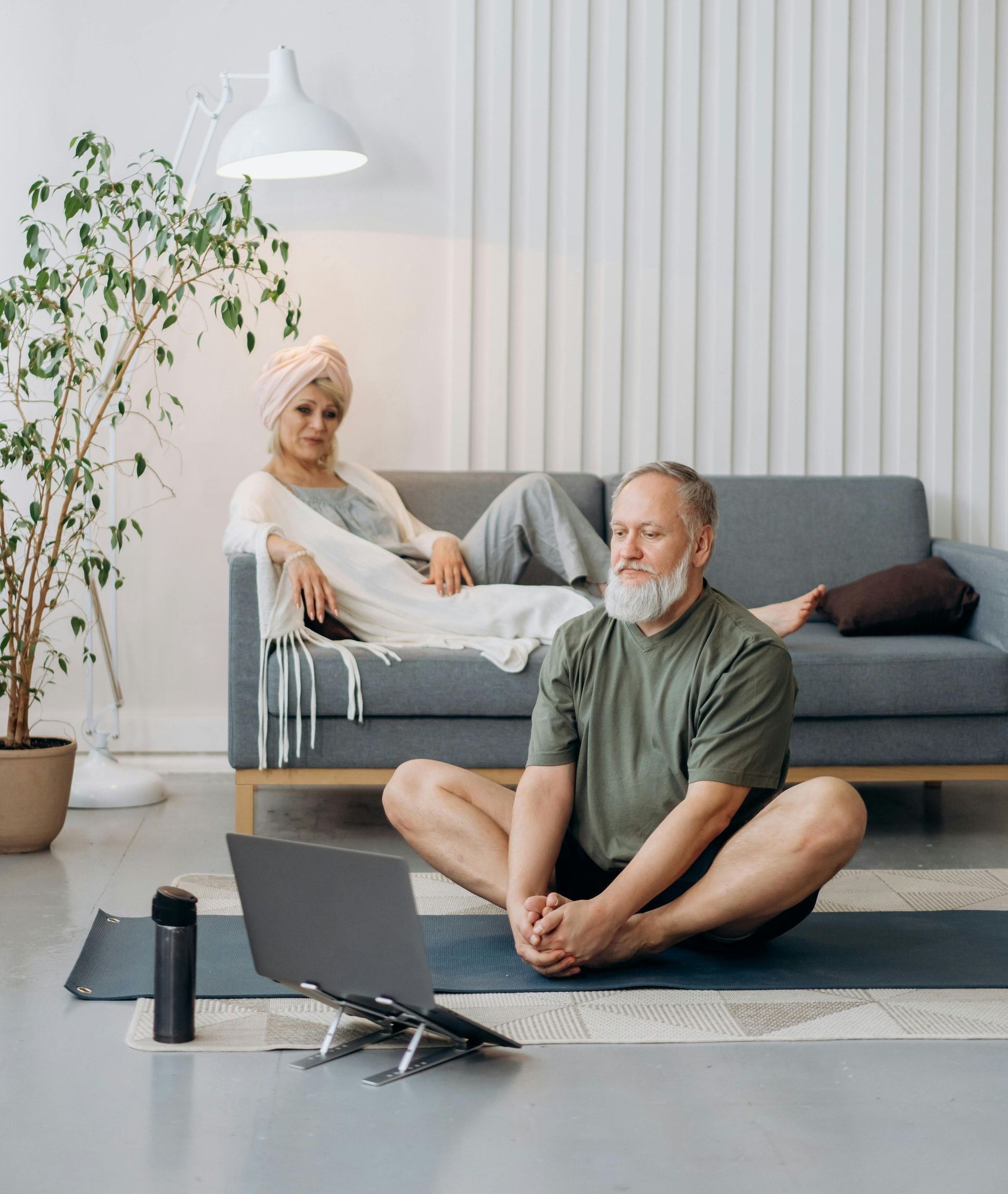 Un homme âgé fait du yoga devant son ordinateur portable, une femme se détend sur un canapé. Intérieur baigné de lumière naturelle.