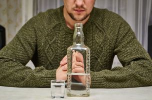 Un homme, les mains jointes, regarde une bouteille et un verre à liqueur remplis d'un liquide clair posés sur une table. Il porte un pull vert.