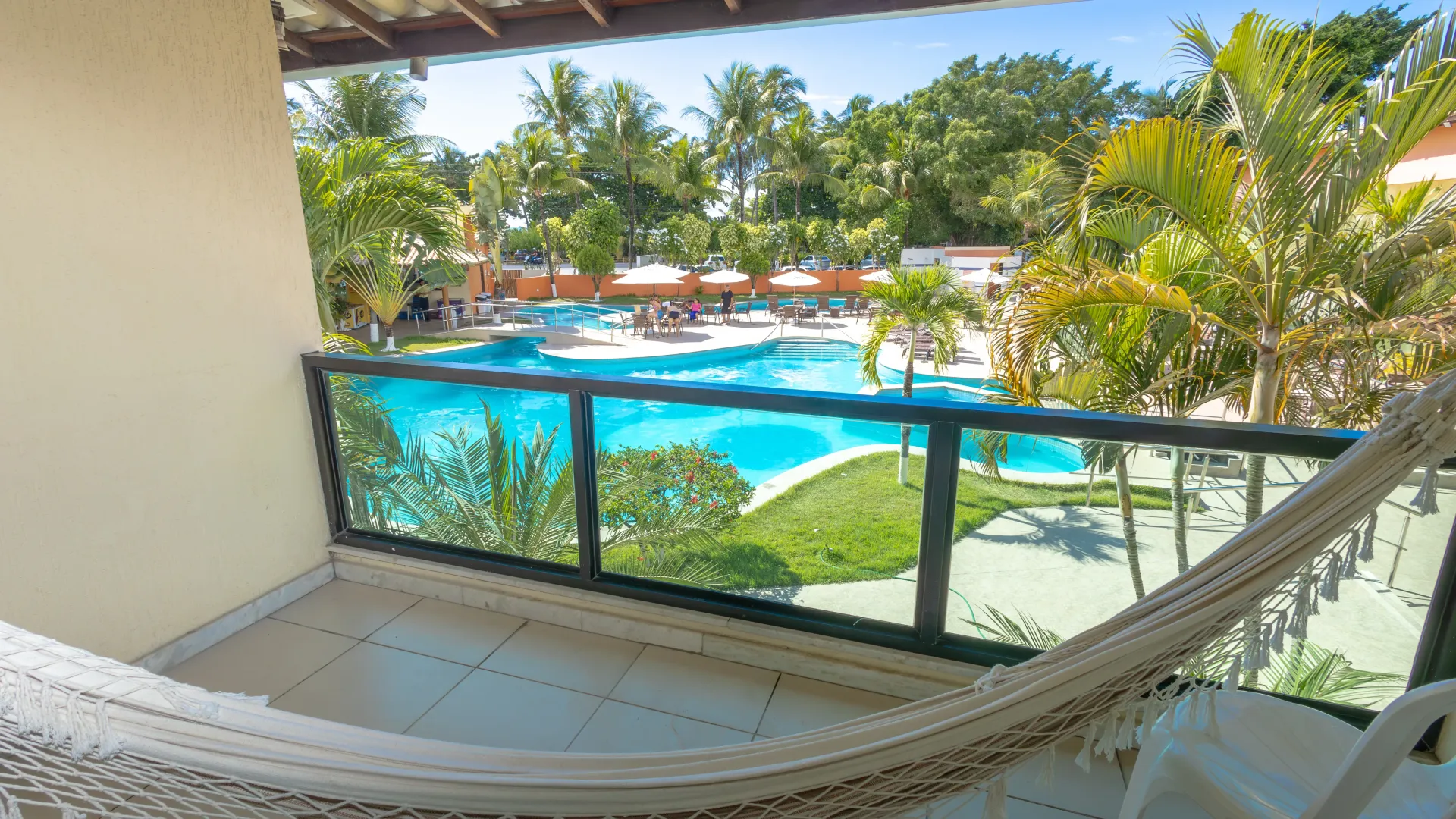 Varanda com vista para a área da piscina, que inclui uma rede, palmeiras e água azul.