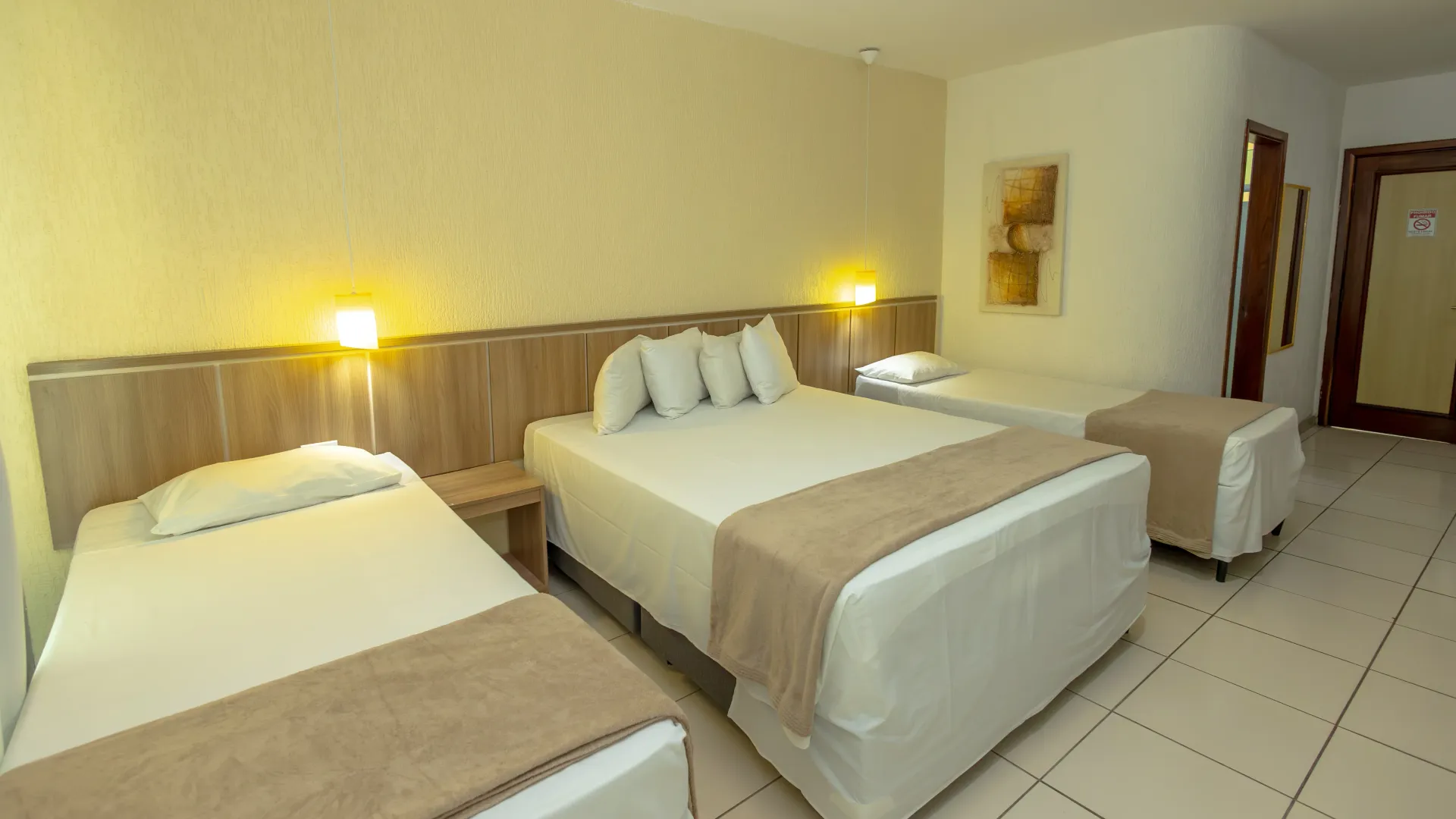 Quarto de hotel com três camas, uma mesa de cabeceira e uma porta, em tons neutros.