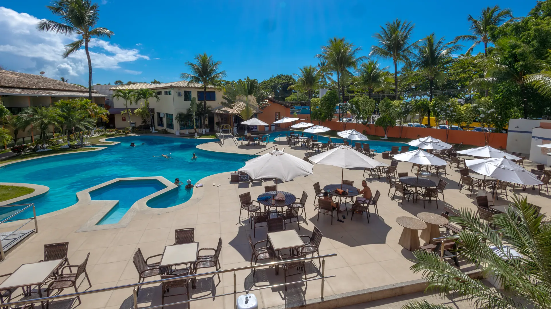 Vista aérea de um resort com edifícios, piscina e guarda-sóis. Árvores verdes rodeiam as estruturas. Hotel Fênix