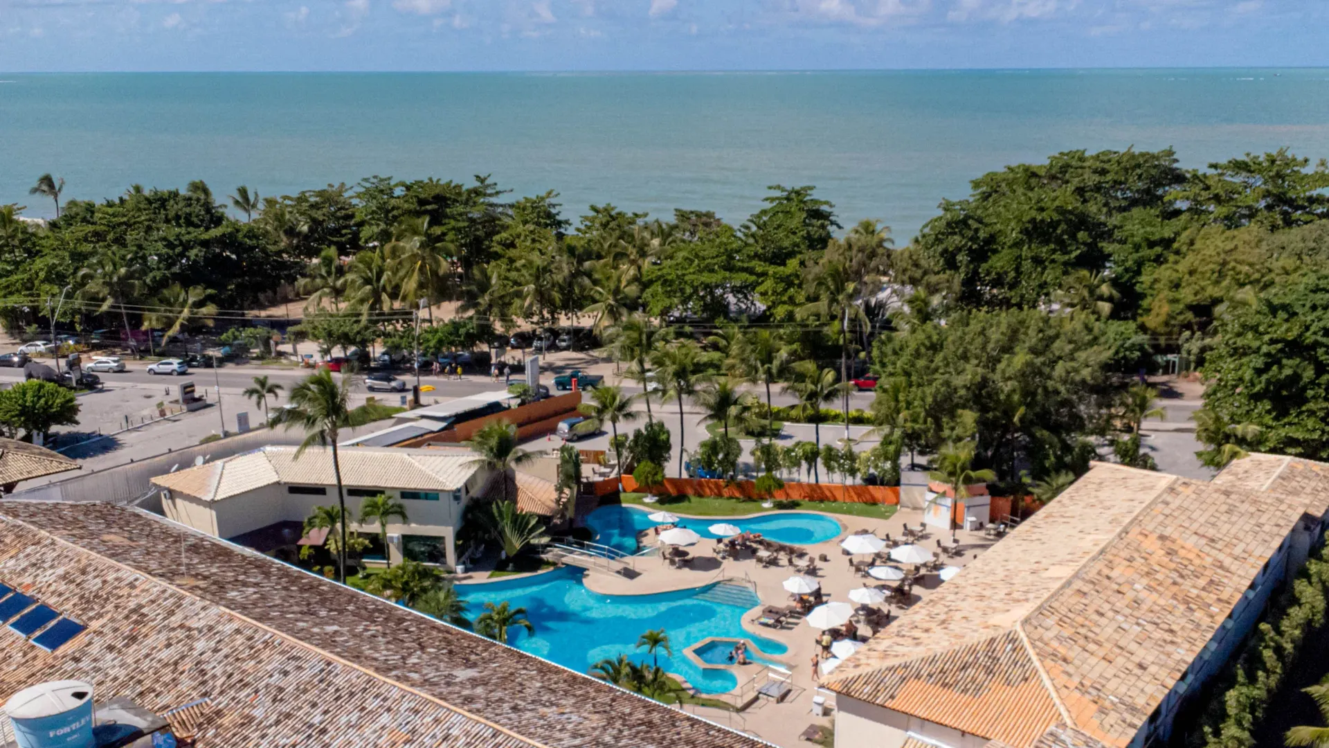 Resort com vista para o oceano, com piscinas, árvores e edifícios, sob um céu azul.
