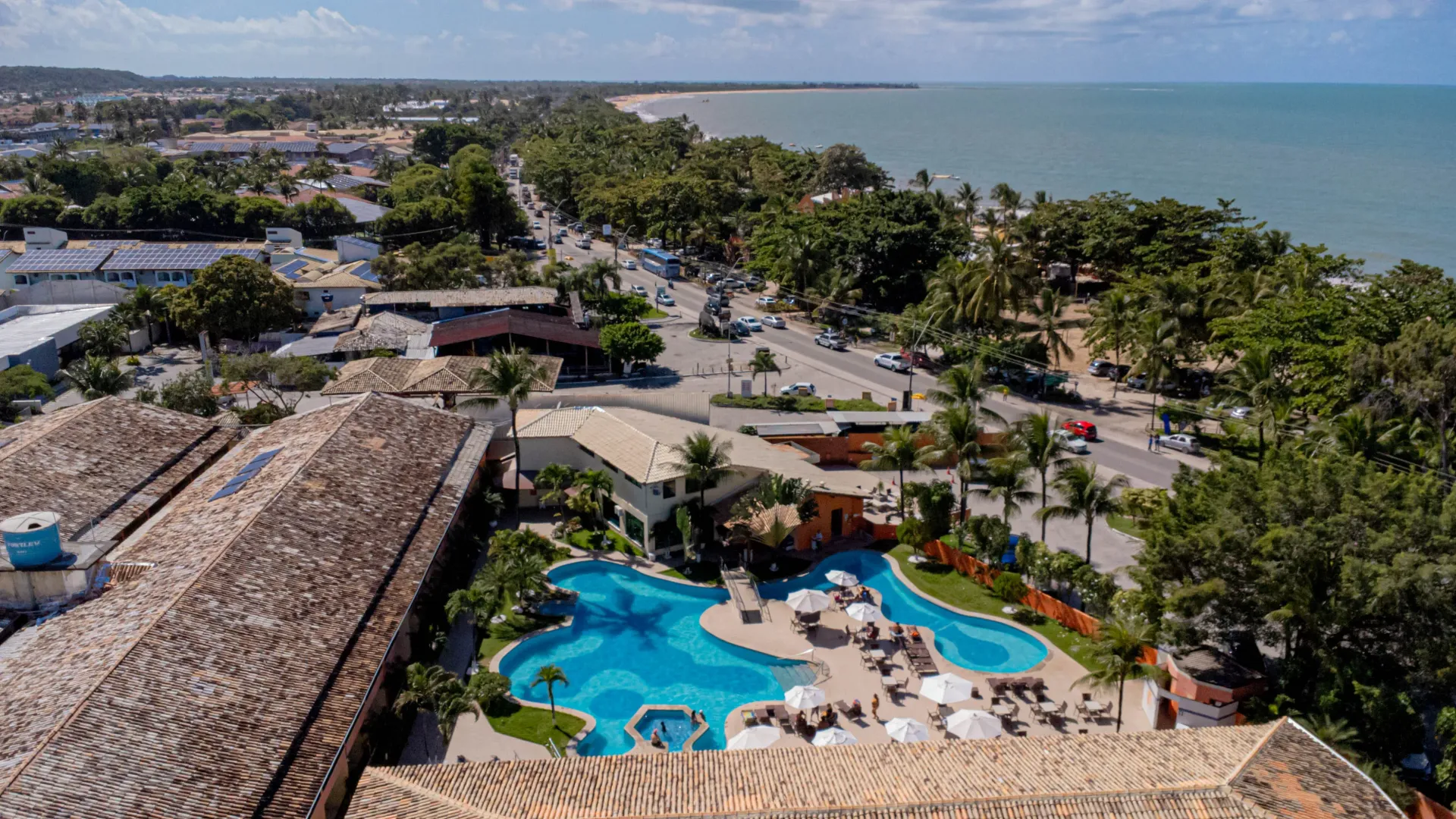 Exterior do hotel com piscina, mesas, guarda-sóis e palmeiras sob um céu azul. Hotel Fênix