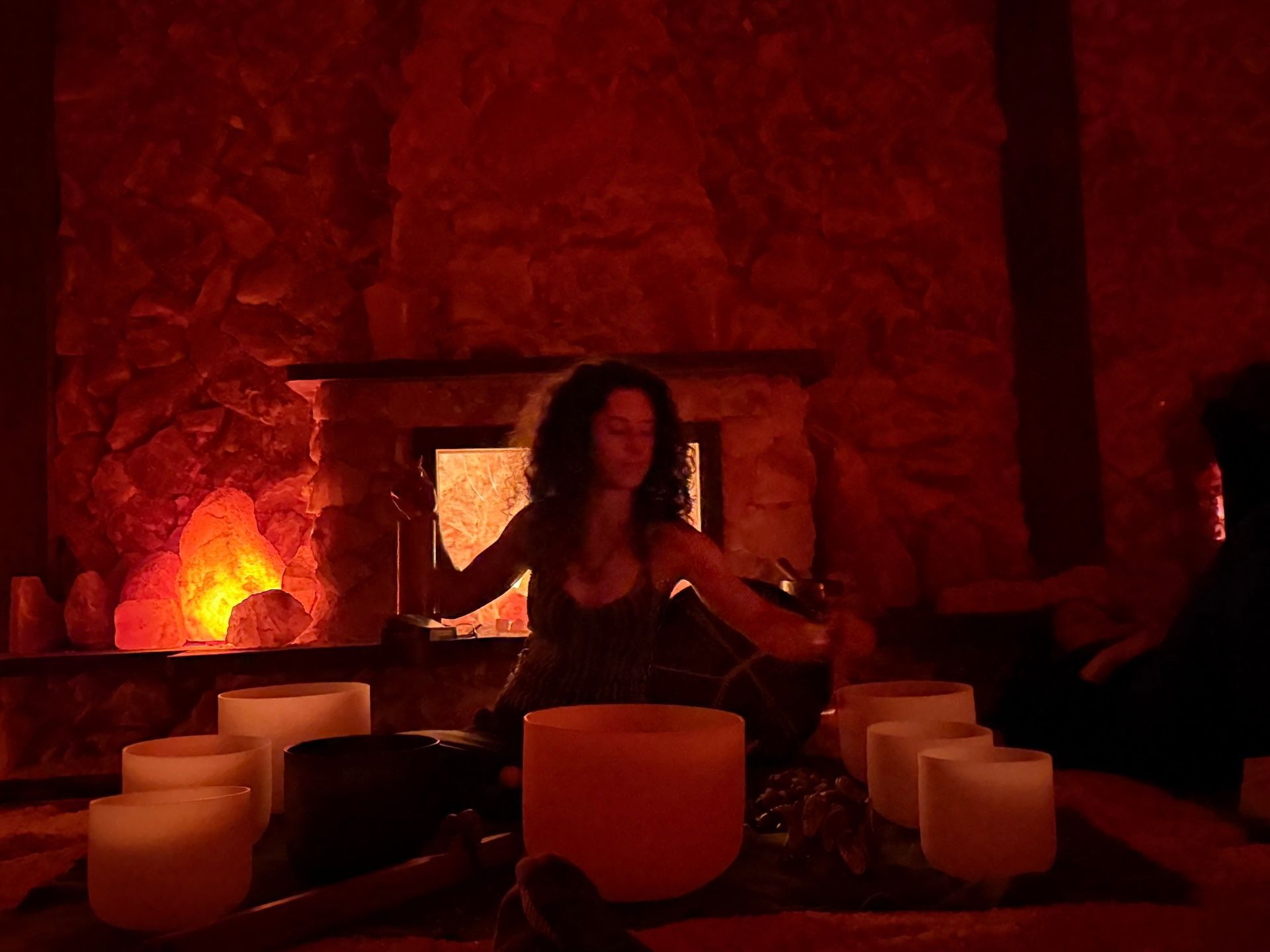 Woman playing crystal bowls in dimly lit room, with fireplace and candles.