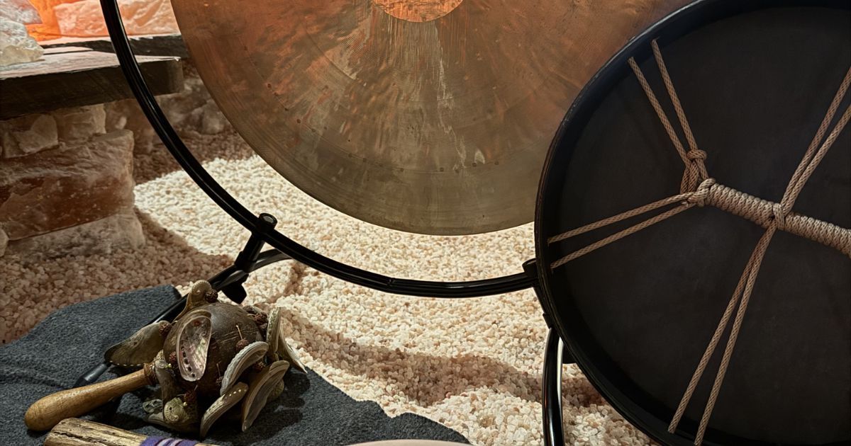 A large metallic gong and a drum with a rope-laced frame stand on a floor covered with light-colored, textured material.