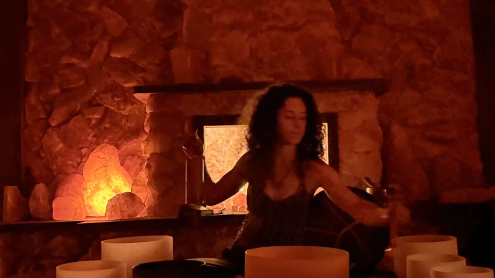 A woman is sitting in front of a fireplace playing a sound bowl.