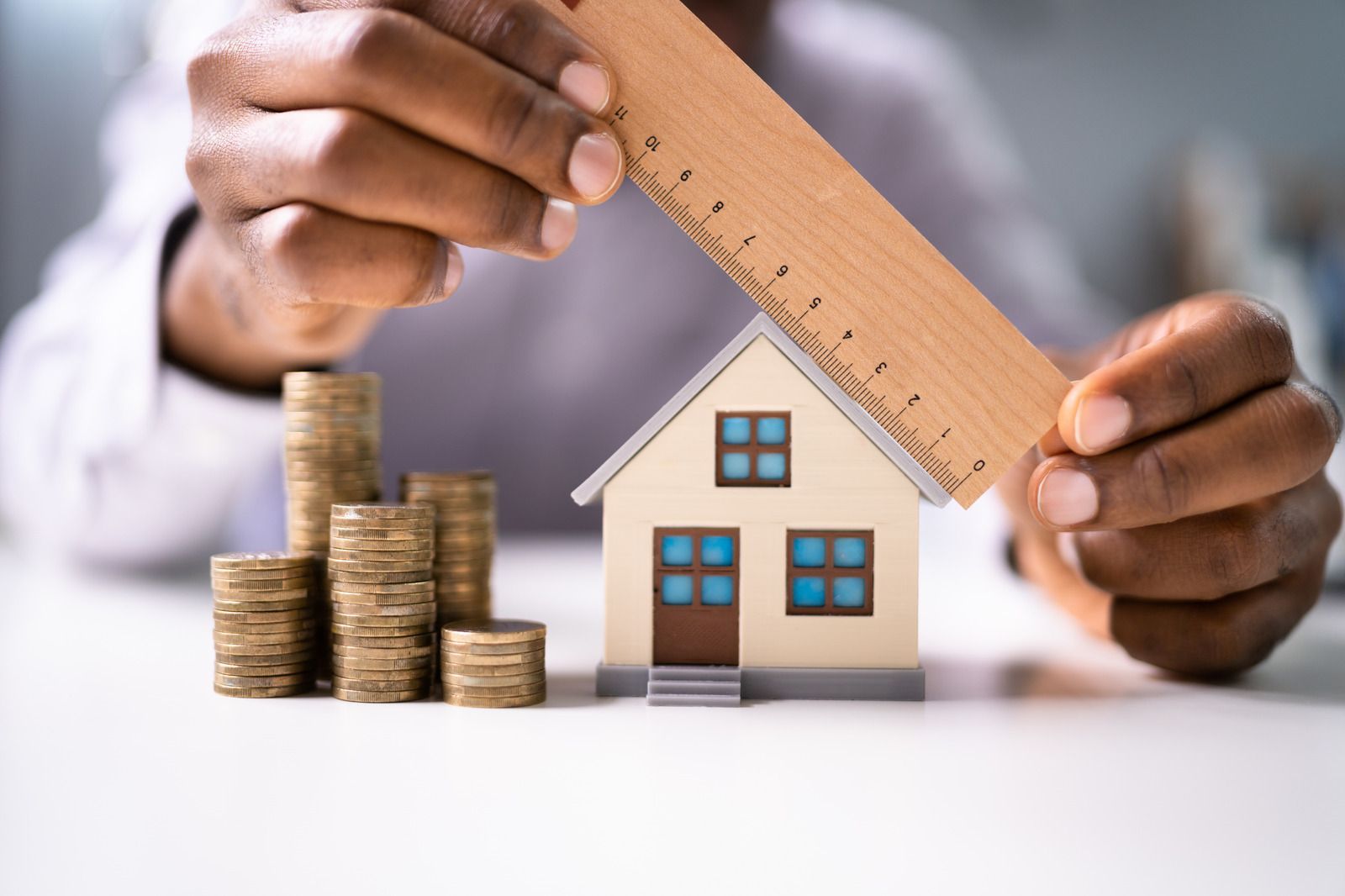A person is holding a ruler over a model house and stacks of coins.