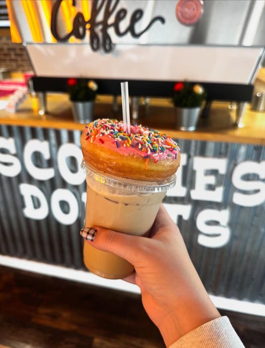 Hand holding iced coffee with a pink frosted donut and sprinkles on top, in front of 
