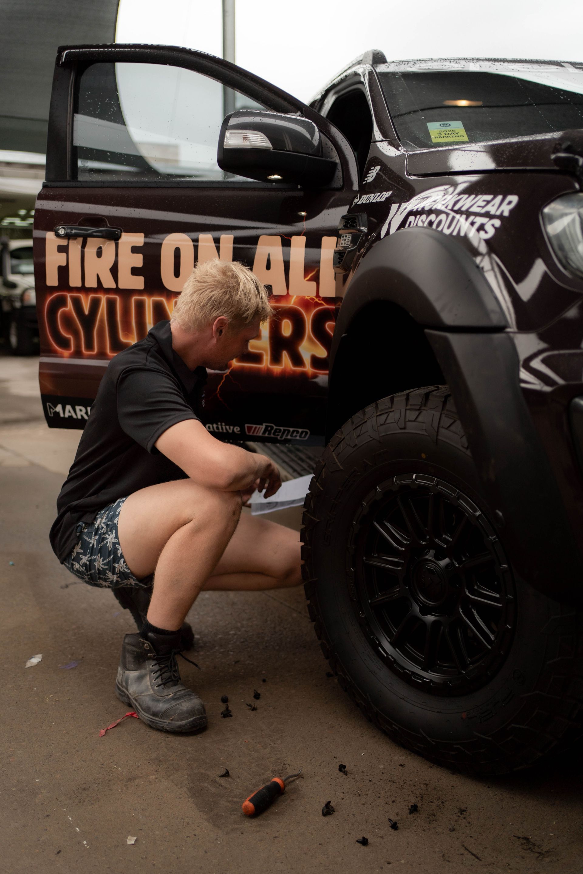 A Man kneeling looking behind front wheels of black car — Douglas Collision Management In Currajong, QLD