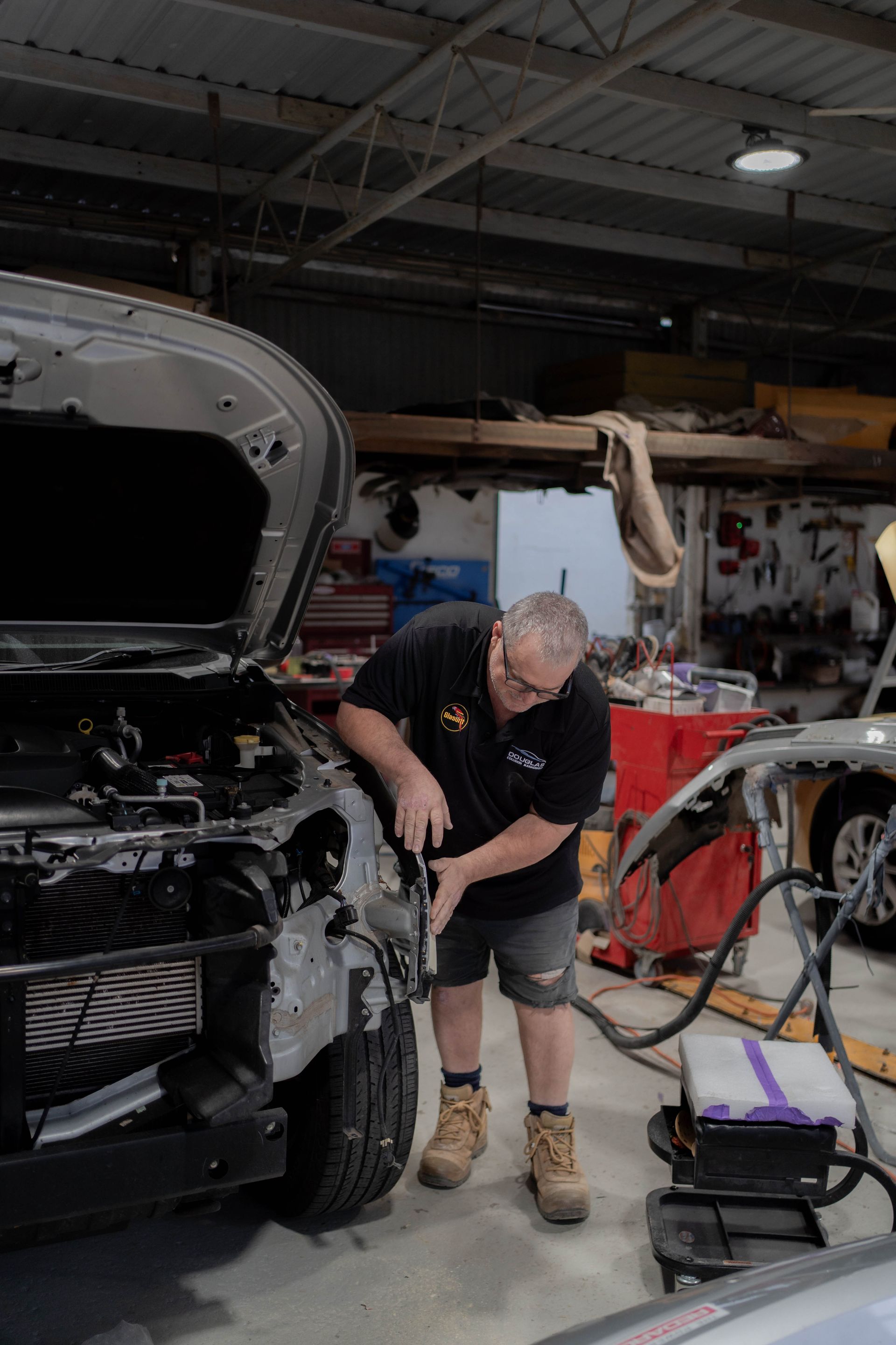Mechanic working on a car in a workshop, using tools. Silver car, dark shirt, shorts — Douglas Collision Management In Currajong, QLD