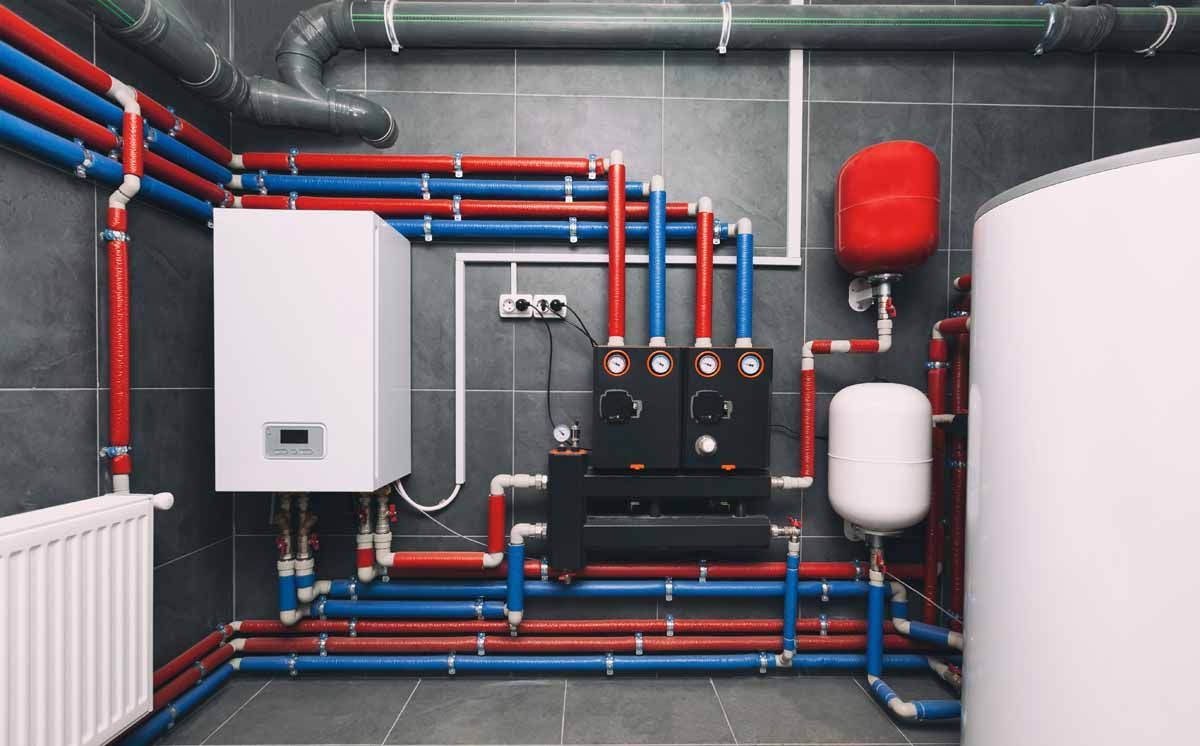 Heating system with red and blue pipes, white tank, and other components against a gray tiled wall.
