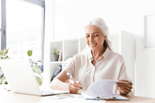 An elderly woman is sitting at a desk with a laptop and a pen.