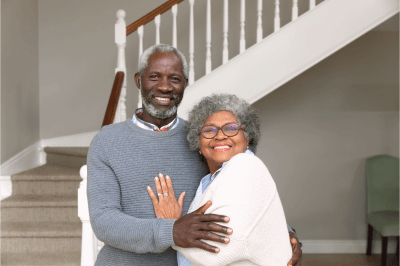 An elderly couple is hugging each other in front of a staircase.