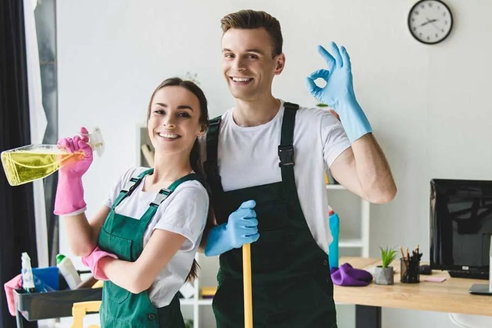 A man and a woman are standing next to each other in an office holding cleaning supplies.