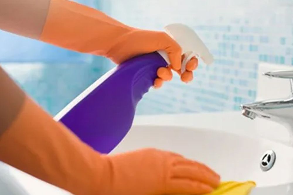 A person wearing orange gloves is cleaning a bathroom sink with a spray bottle.