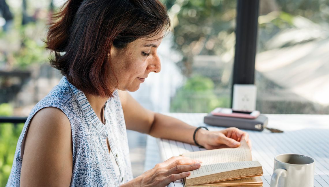 Mujer sentada junto a una ventana mientras lee un libro en una mesa con una taza al lado.