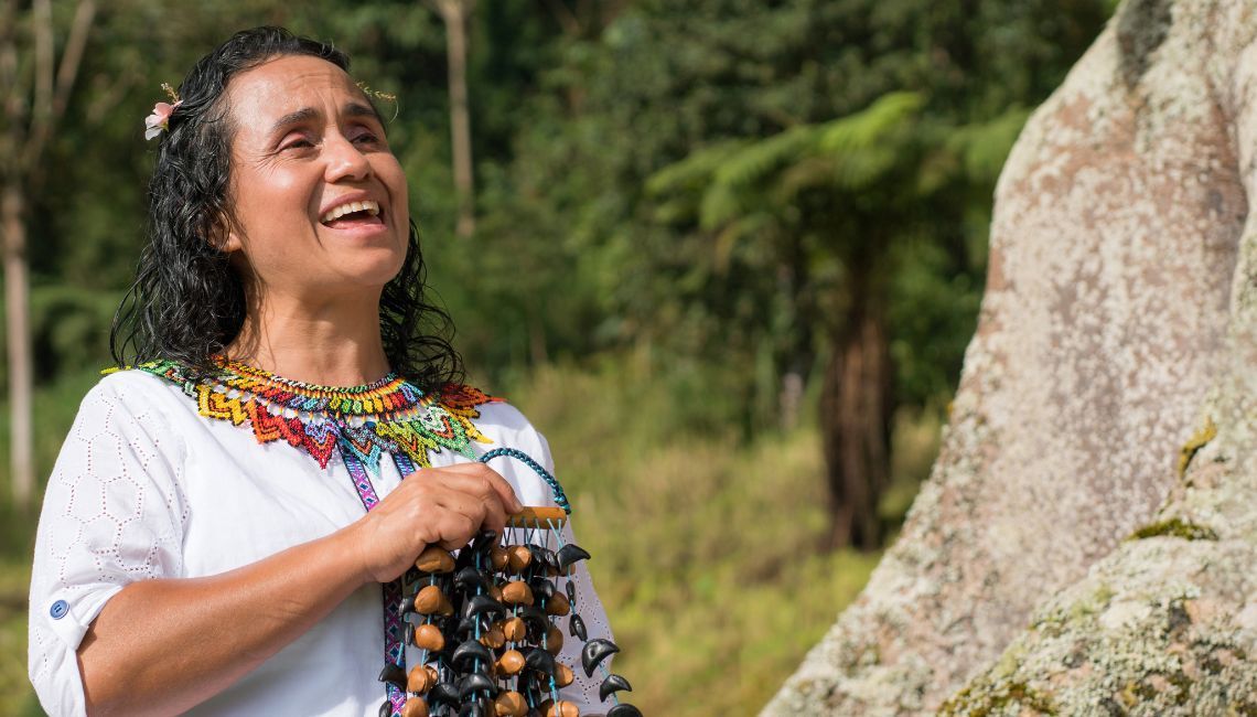 Mujer indígena en ceremonia ritual al aire libre, vestida con bordado tradicional y rodeada de naturaleza, expresión de conexión espiritual con la tierra.