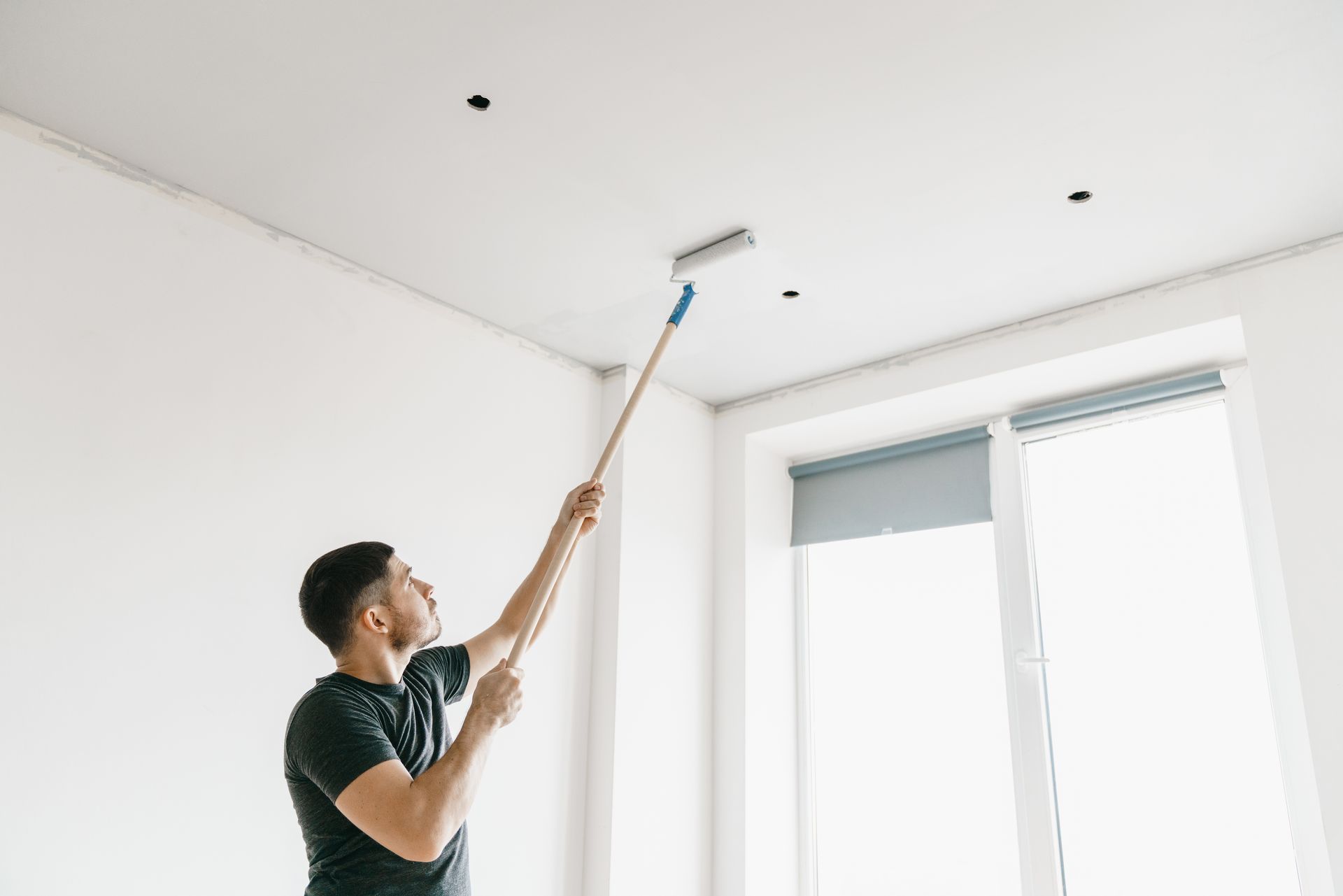 Man painting a white ceiling with a roller, in a room with a window.