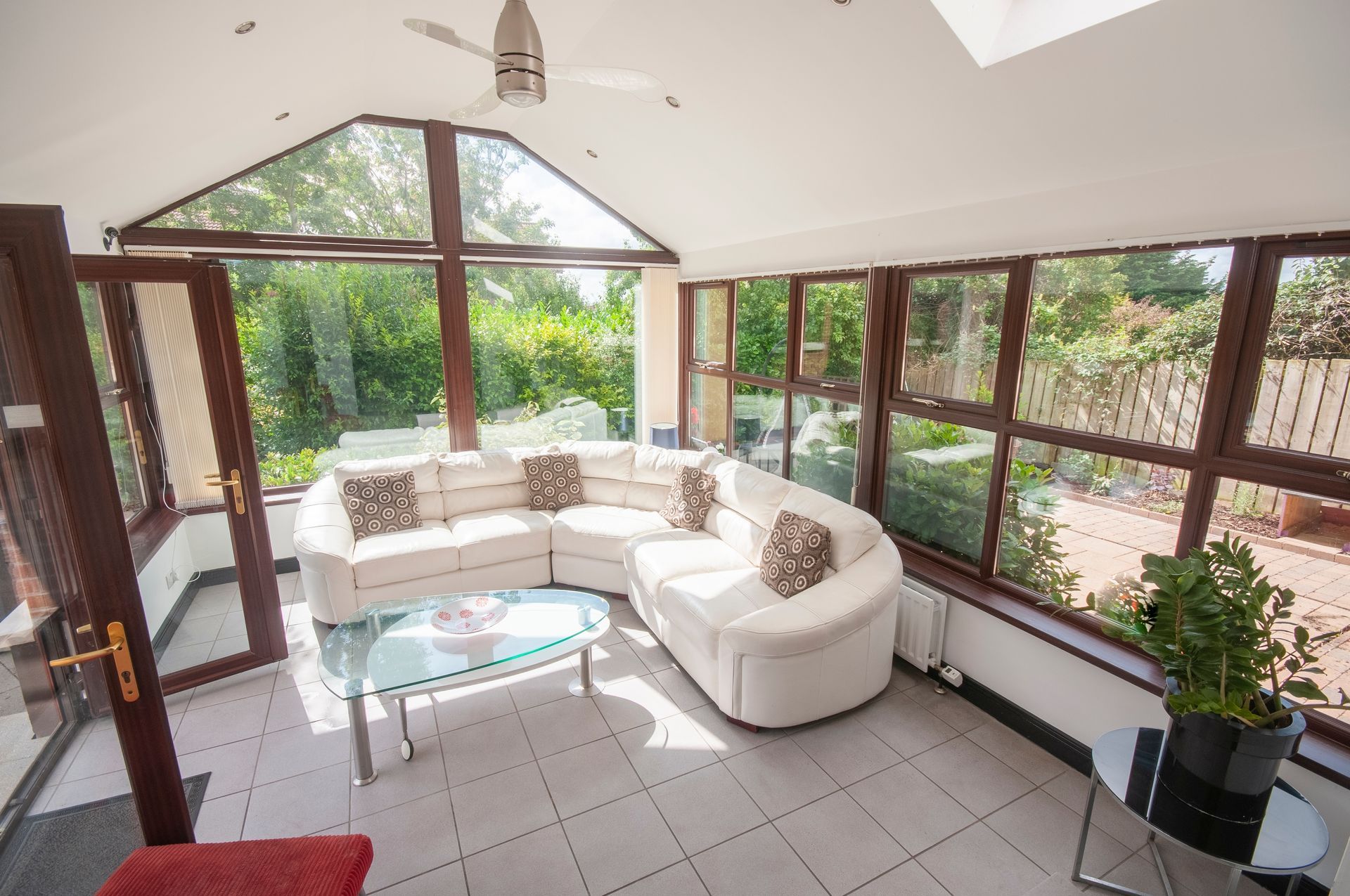 Sunroom with white sectional sofa, glass table, and lush greenery outside windows.
