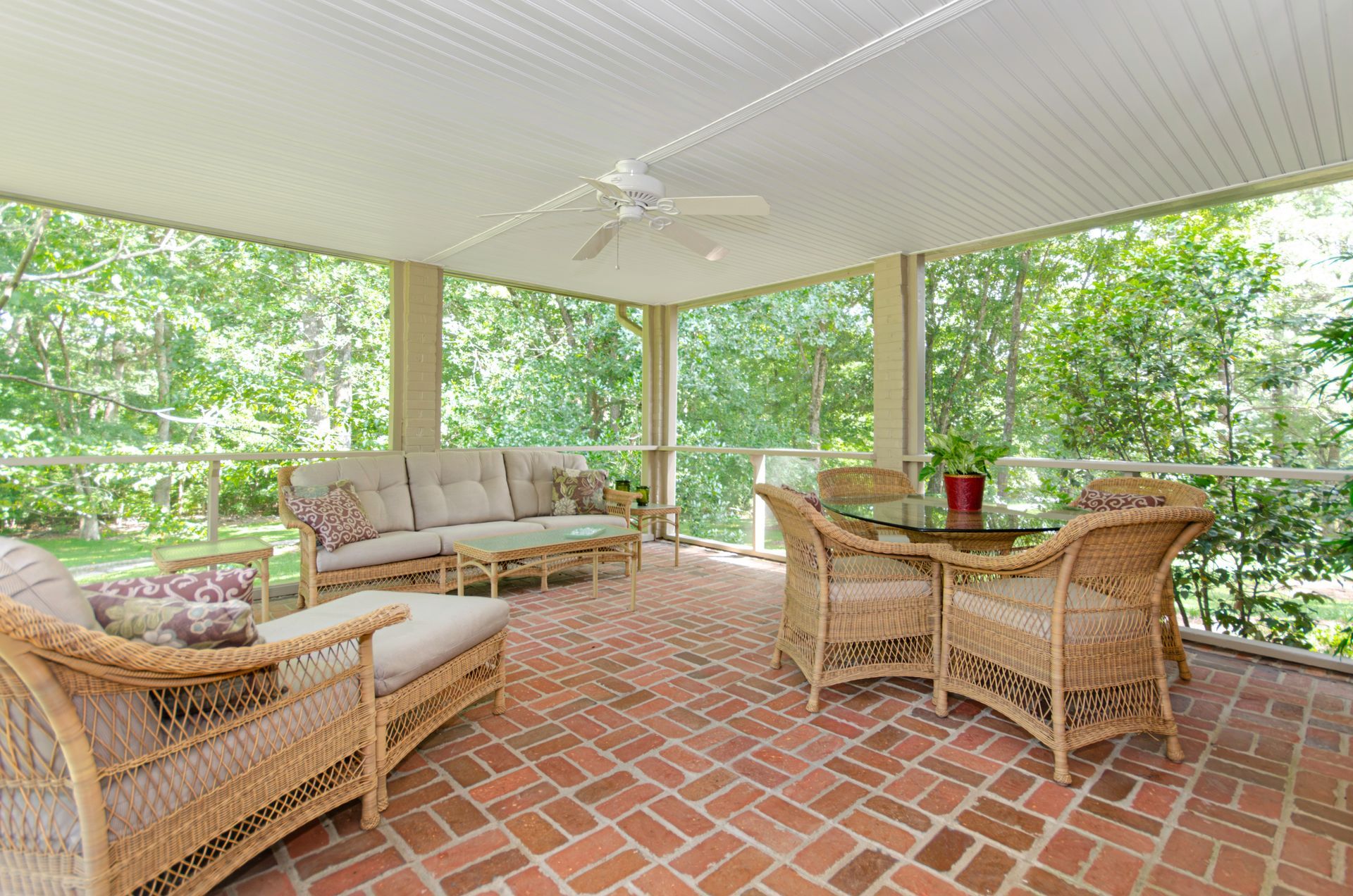 Screened-in patio with brick floor, wicker furniture, and a ceiling fan, overlooking a wooded area.