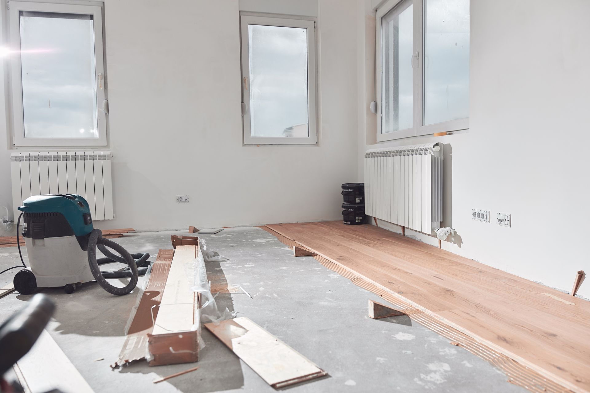 Installing hardwood flooring in a room with white walls, windows, and a vacuum cleaner.