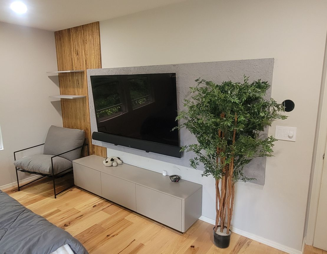 Modern bedroom with mounted TV, soundbar, and entertainment center against gray wall; wood accent and armchair.