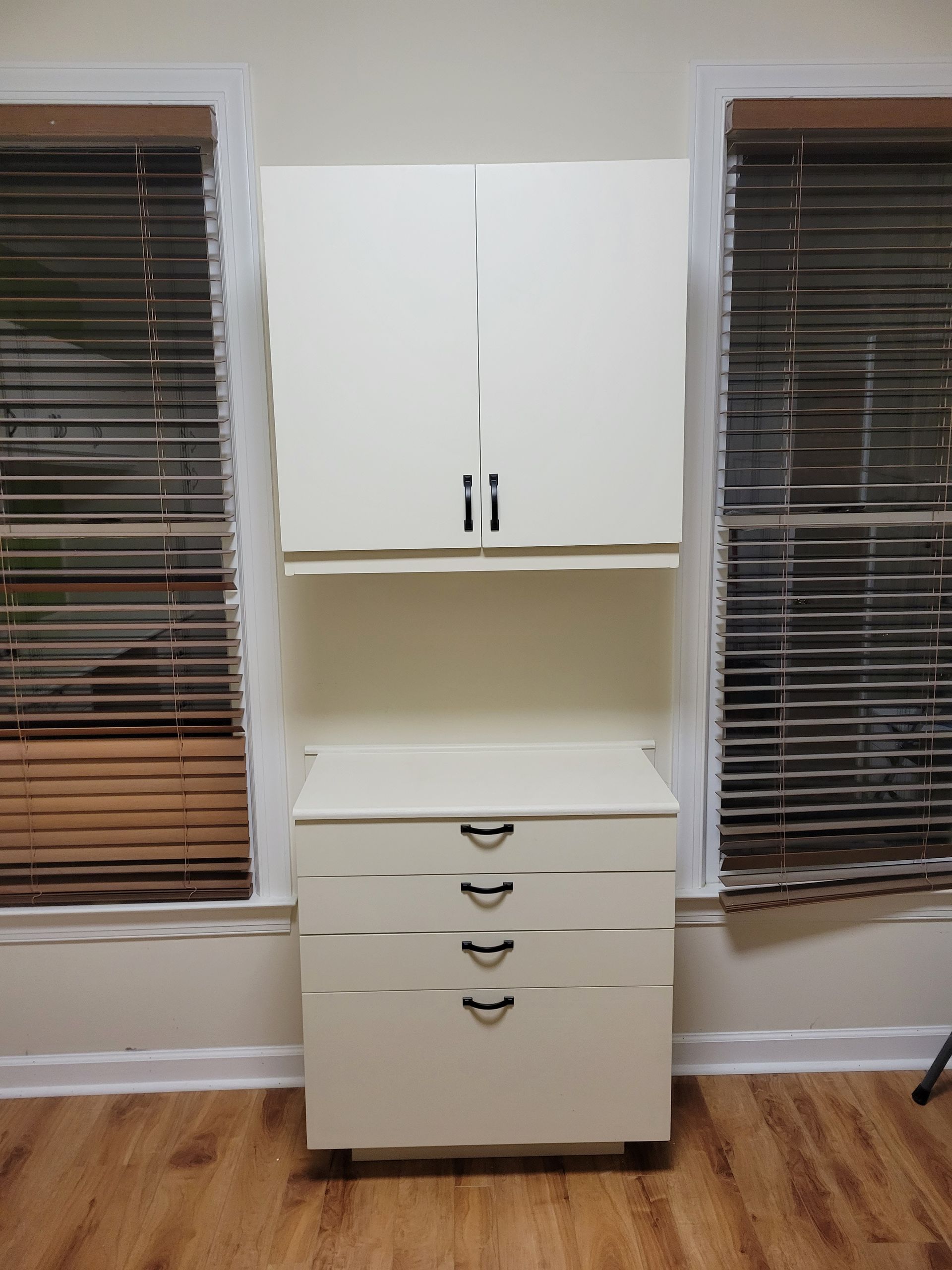 A white cabinet with drawers is in a room next to two windows.