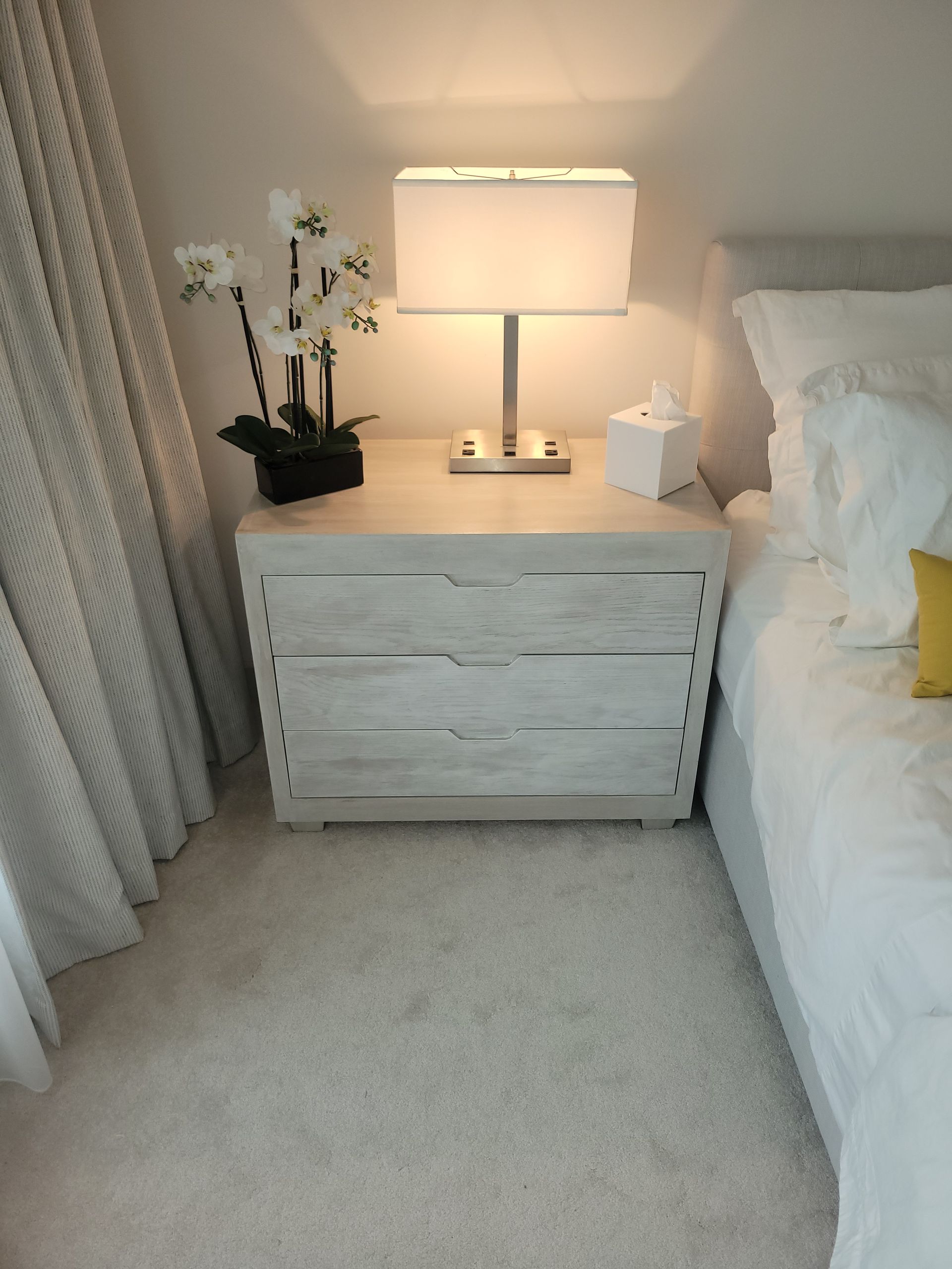 A nightstand with a lamp and flowers on it next to a bed