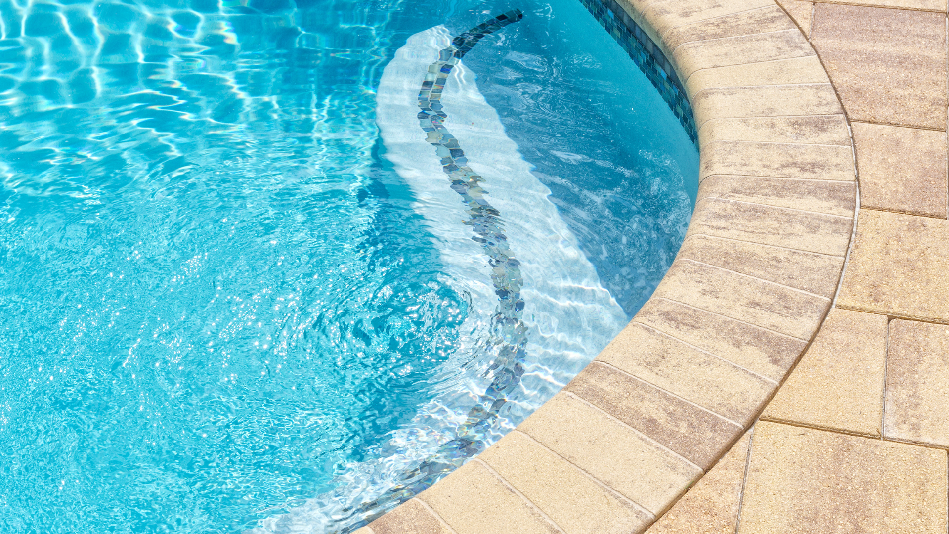 A tablet , hat , sunglasses , and a glass of water are sitting next to a swimming pool.
