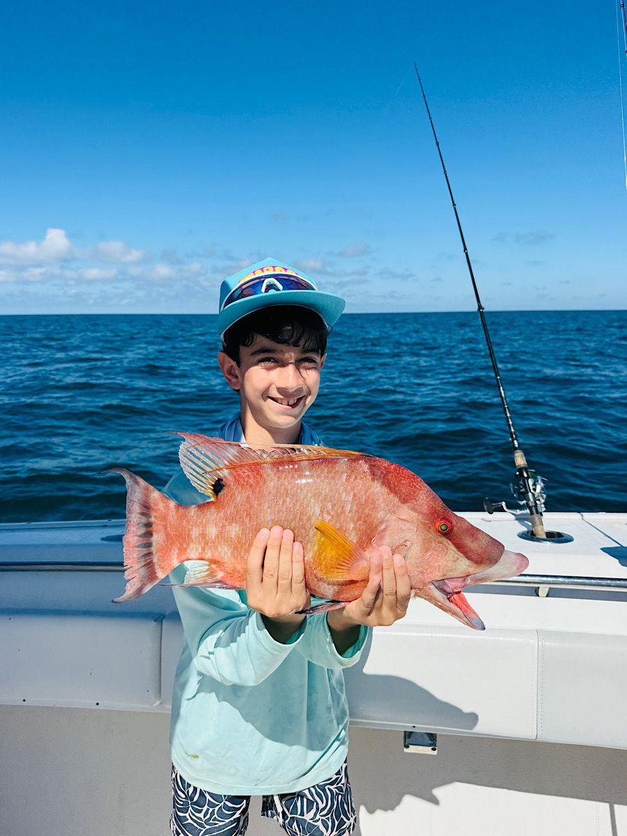 Boy on boat smiles, holding a large, reddish fish, fishing rod in background, ocean.