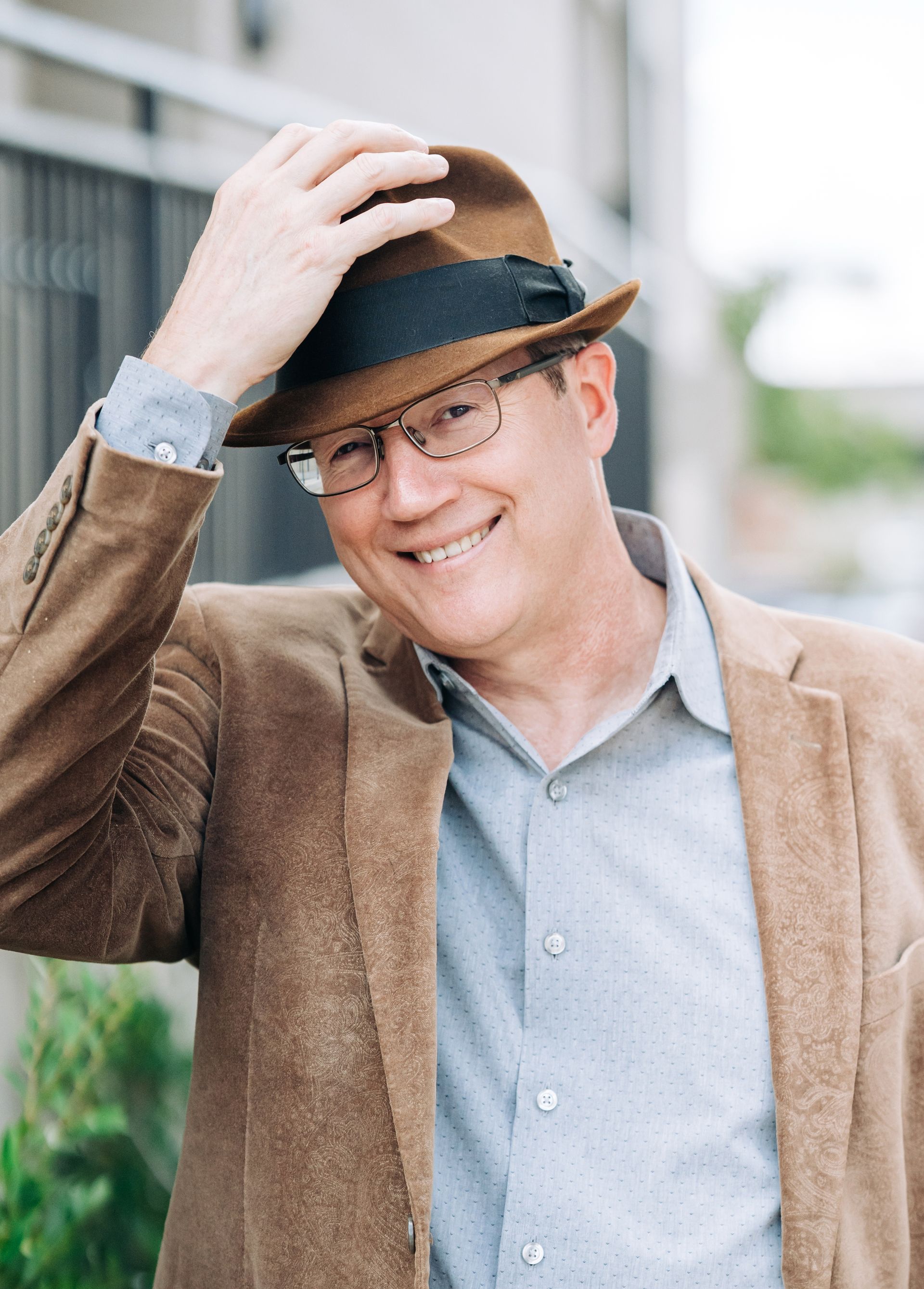 Man wearing glasses, a striped pink shirt, and a navy blue blazer smiling in front of a dark background.