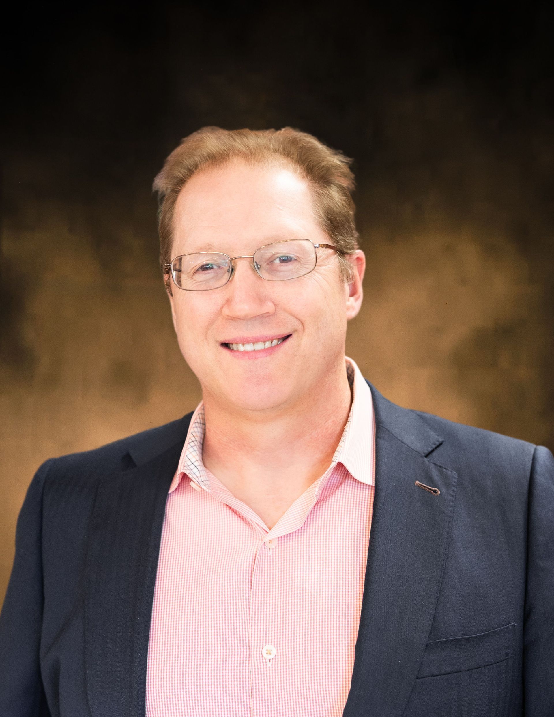 Man wearing glasses, a striped pink shirt, and a navy blue blazer smiling in front of a dark background.