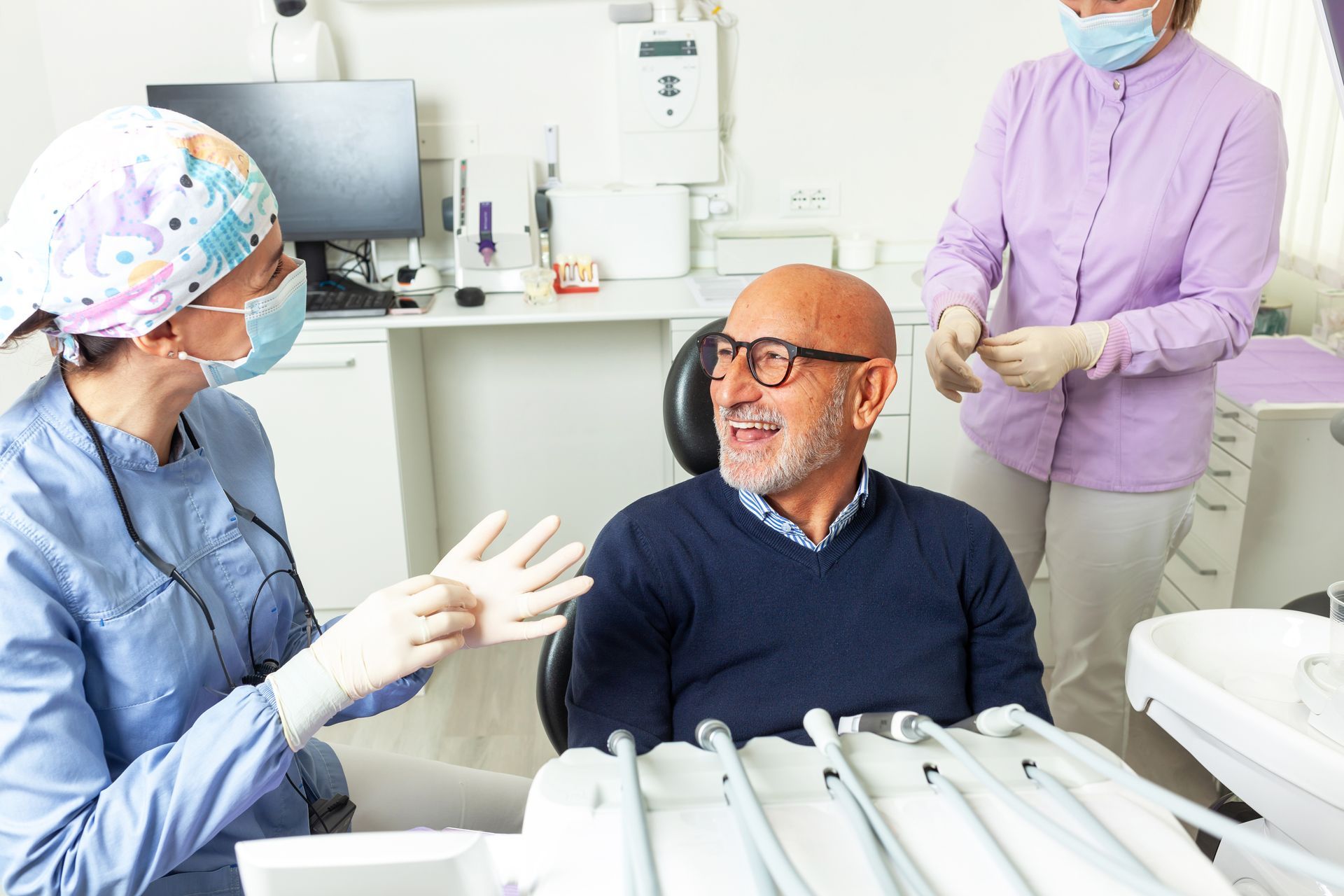 Dentist in scrubs and face mask talks to a patient in a dental chair; assistant stands nearby.