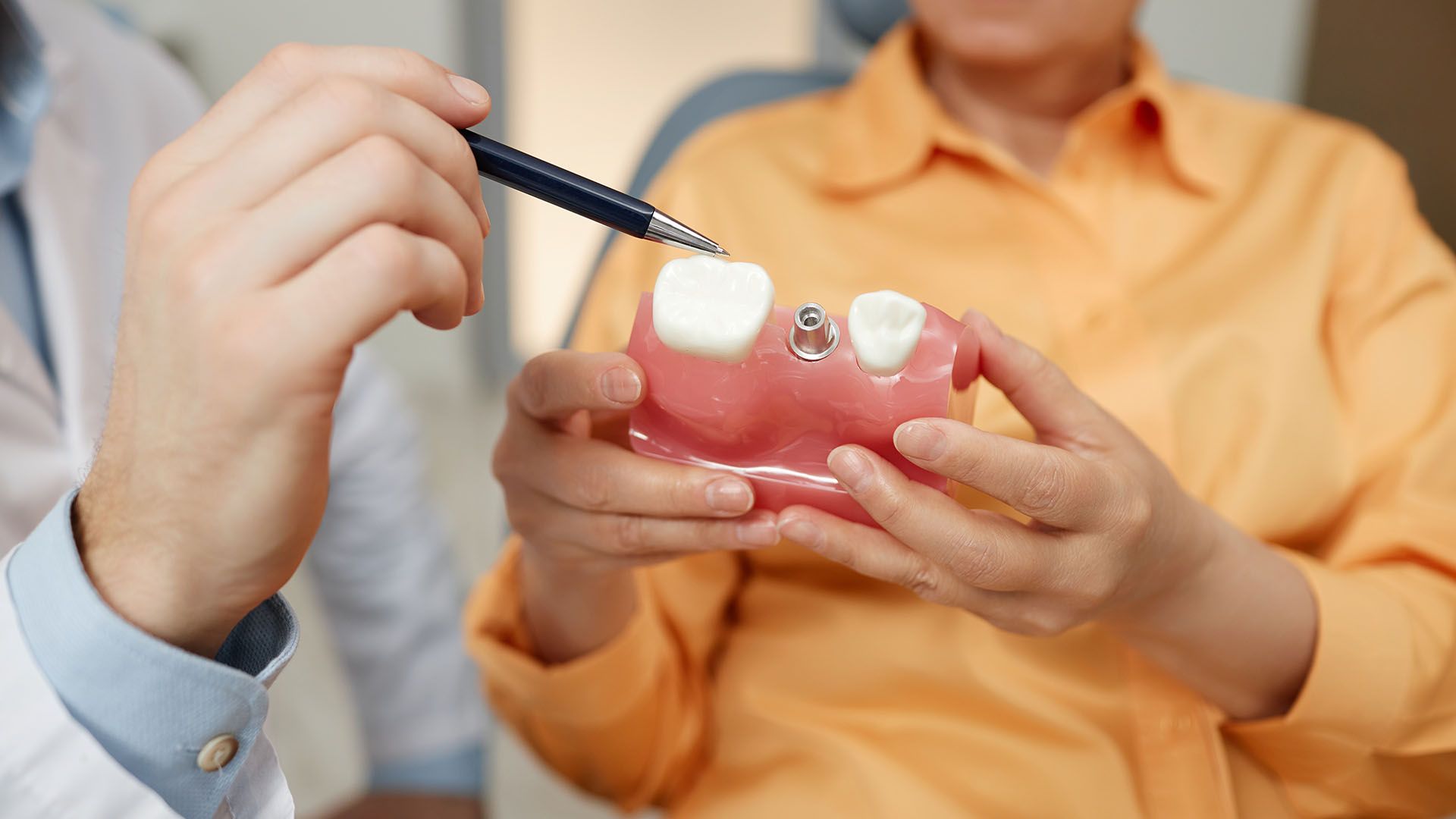 Dentist pointing to dental implant model held by a patient in an orange shirt.