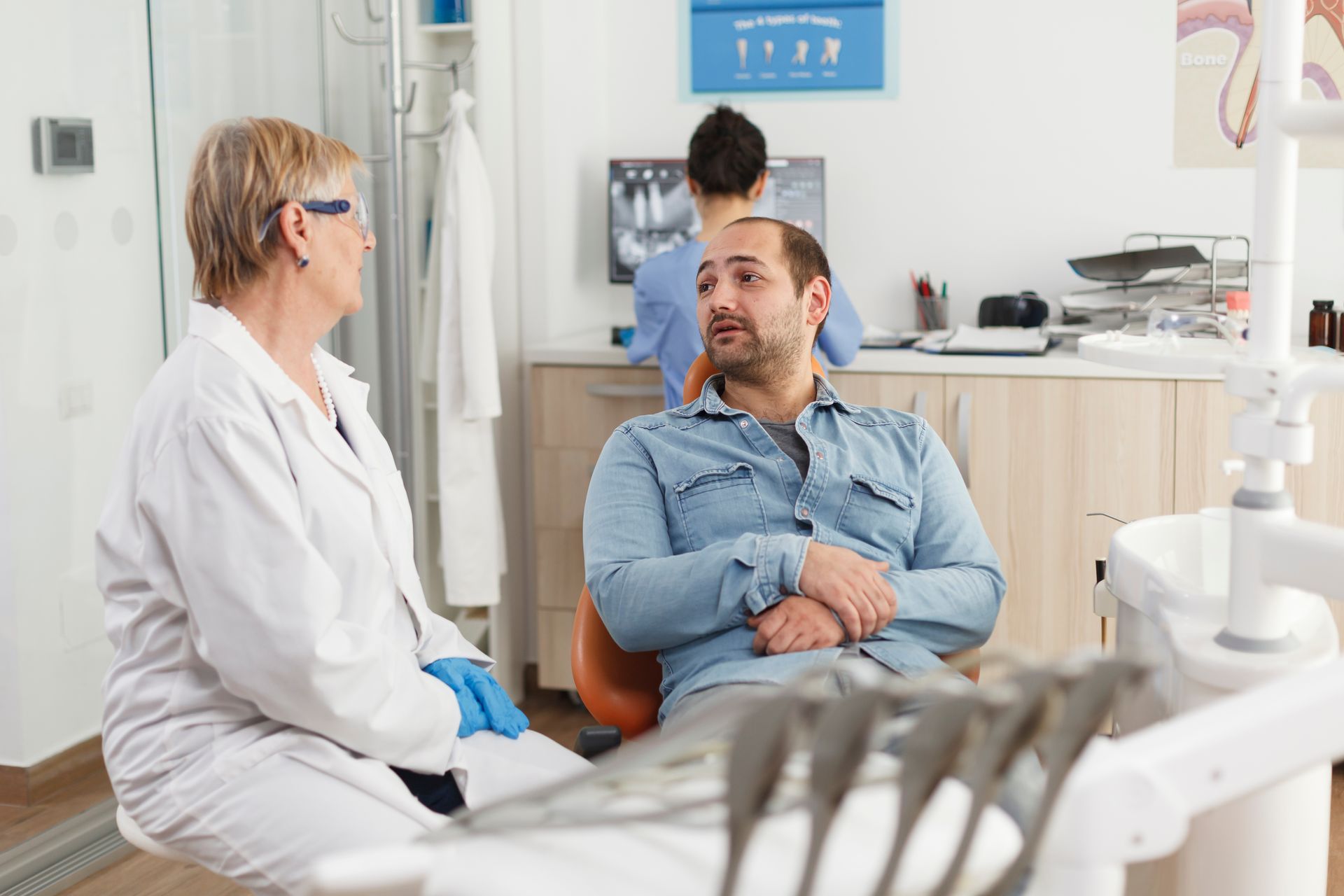 Dentist talking to a patient in an examination room; assistant behind them.