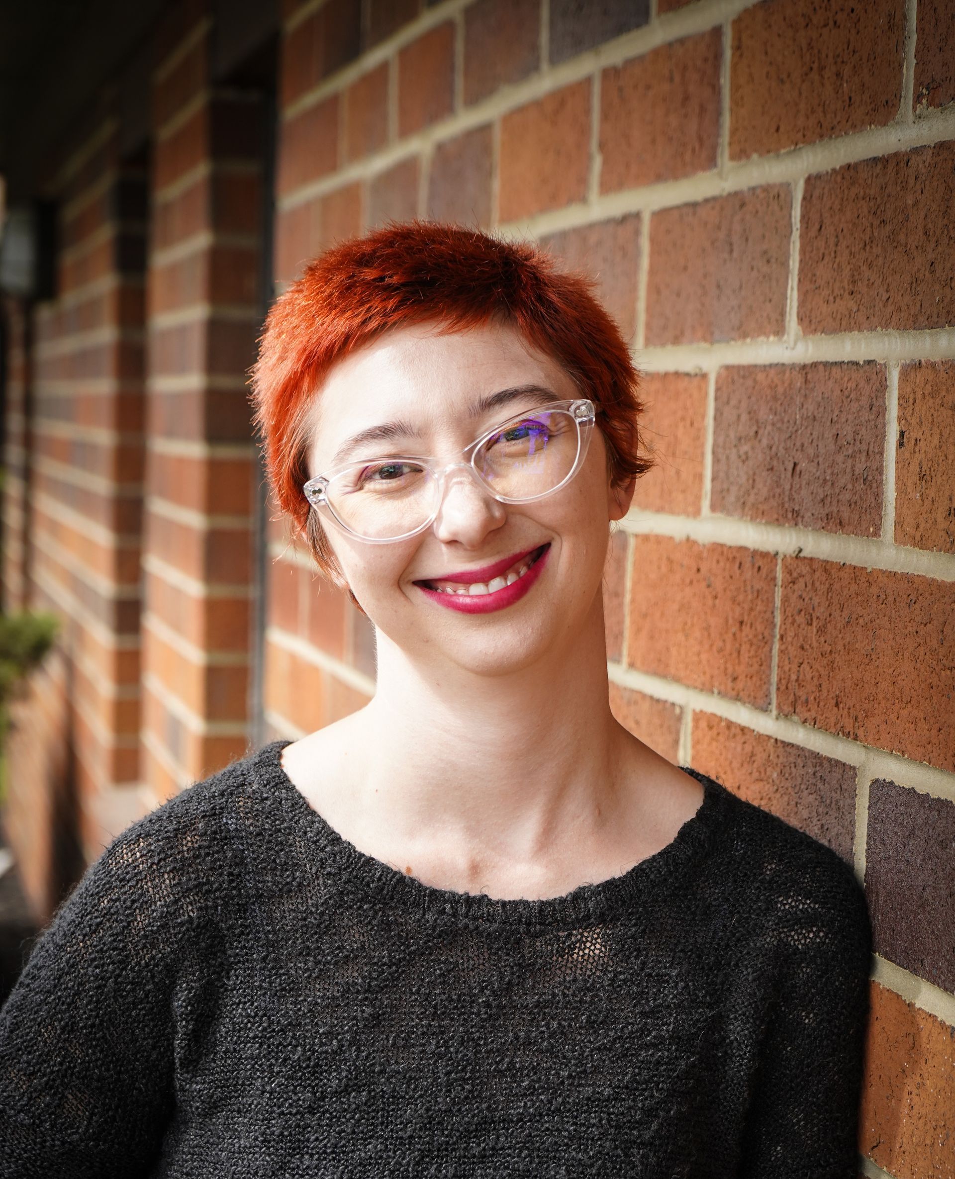A woman with red hair and glasses is smiling in front of a brick wall.