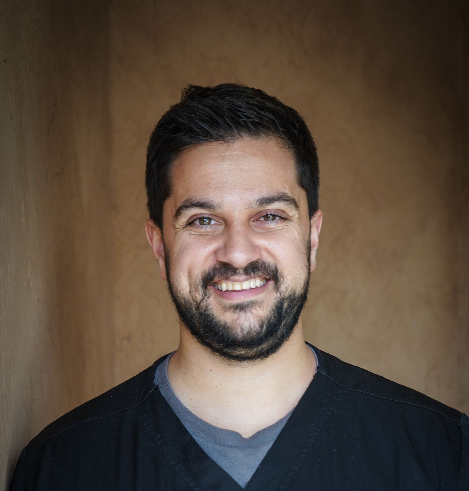A man with a beard is smiling for the camera while wearing a black scrub top.