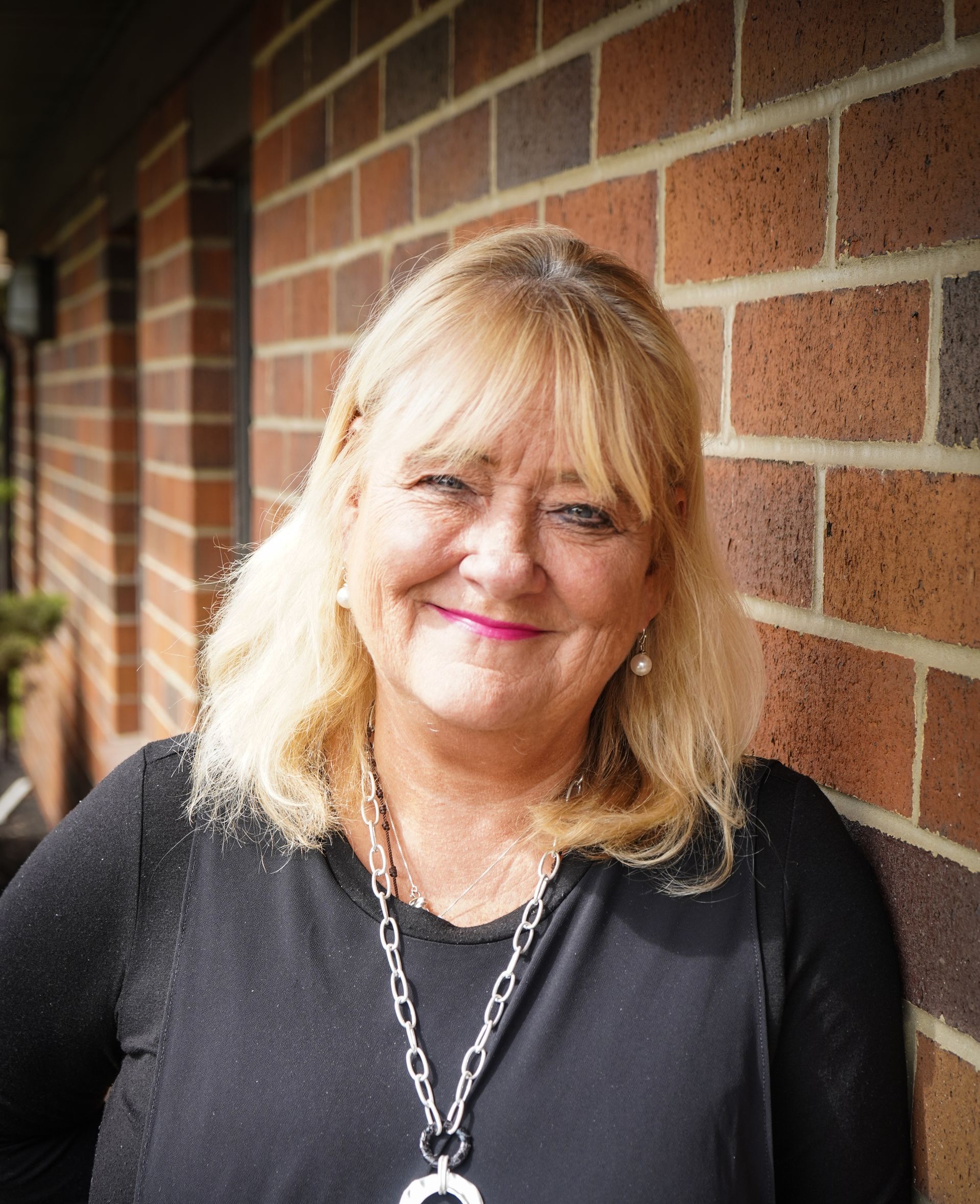 A woman wearing a black shirt and a necklace is standing in front of a brick wall.