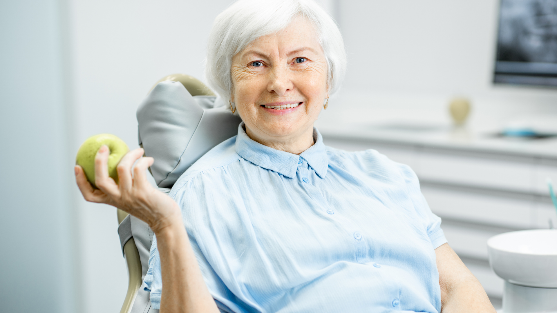 An elderly woman is sitting in a dental chair holding an apple.