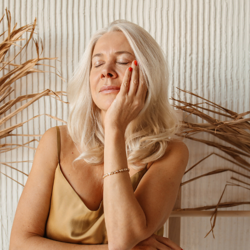 A woman with blonde hair is sitting with her eyes closed and her hand on her face.