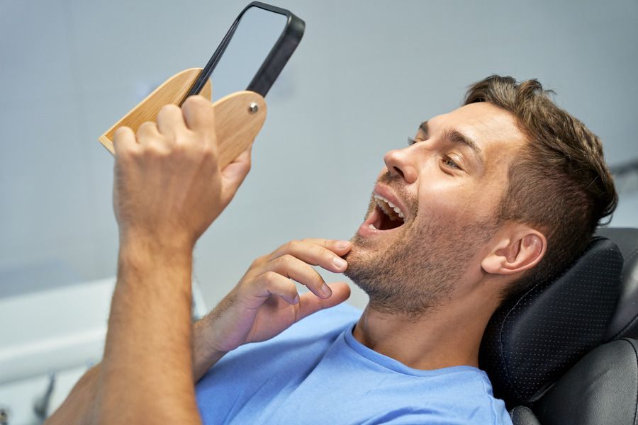 A man is sitting in a dental chair looking at his teeth in a mirror.