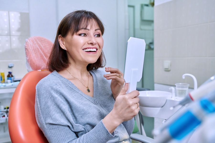 A woman is sitting in a dental chair looking at her teeth in a mirror.