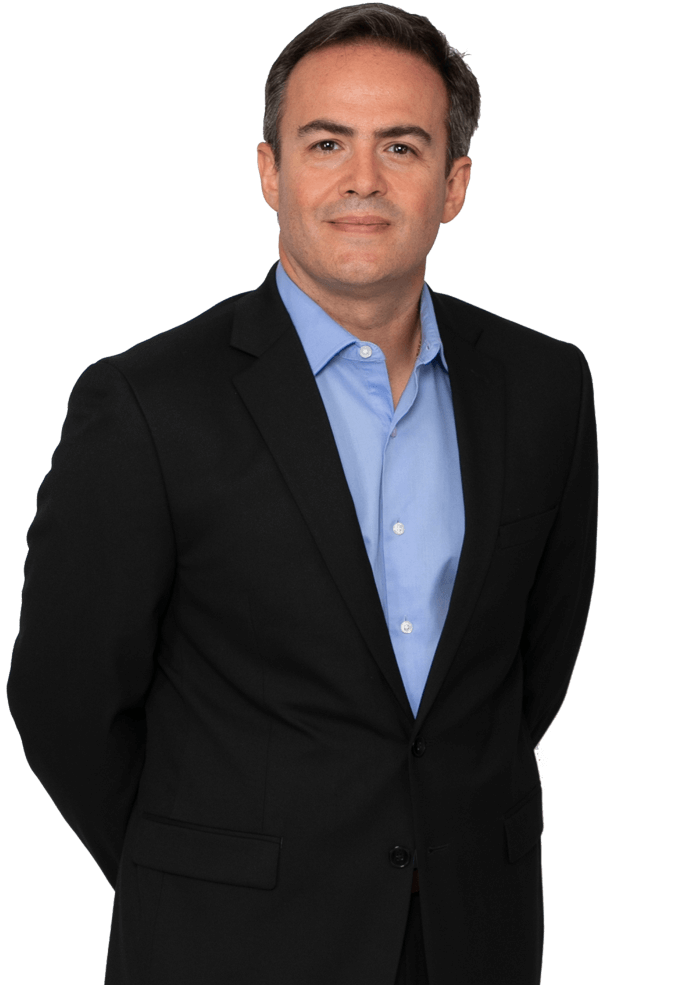 Man in black suit, light blue shirt, hands behind back, smiling.