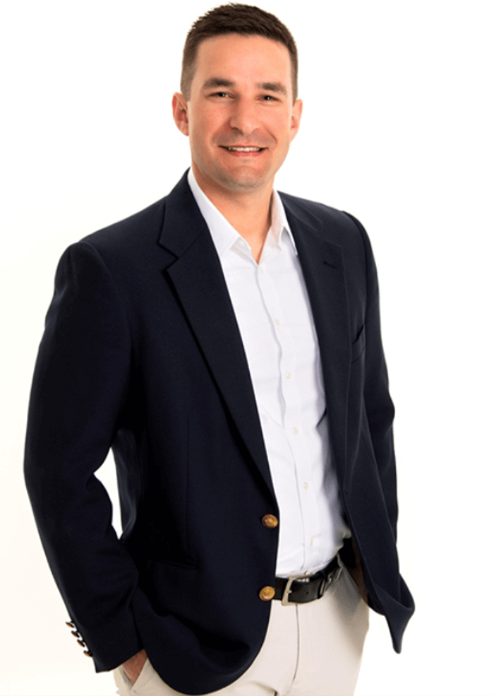 Man in a navy blazer, white shirt, and khaki pants smiles with his hands in his pockets.
