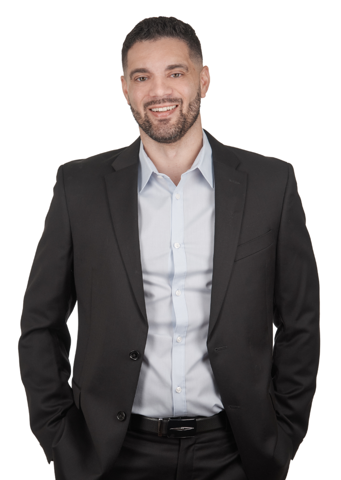 Man in a dark suit smiles, hands in pockets. He wears a light blue shirt.