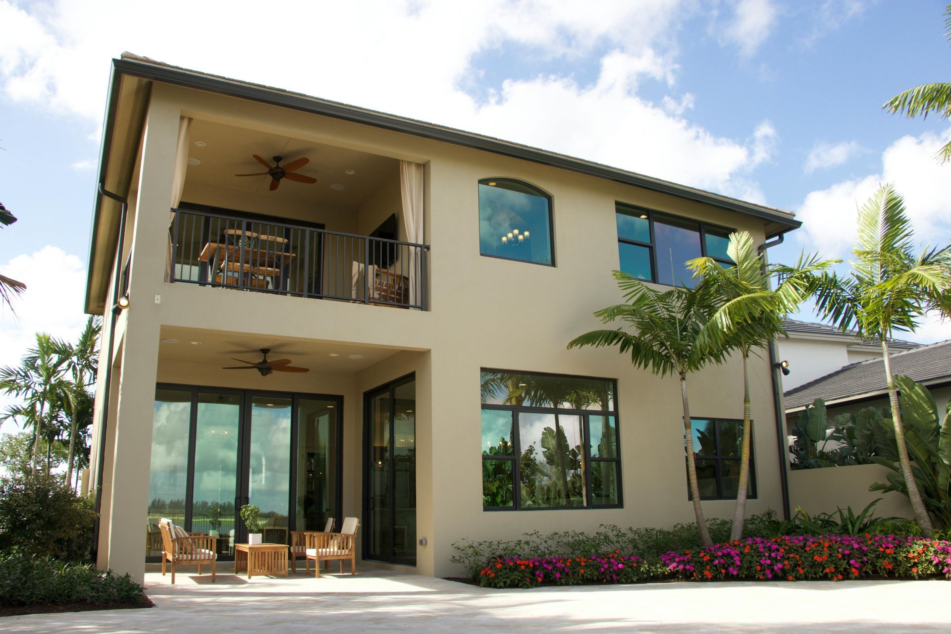 Two-story beige house with balcony, large windows, and outdoor seating. Palm trees and blue sky.