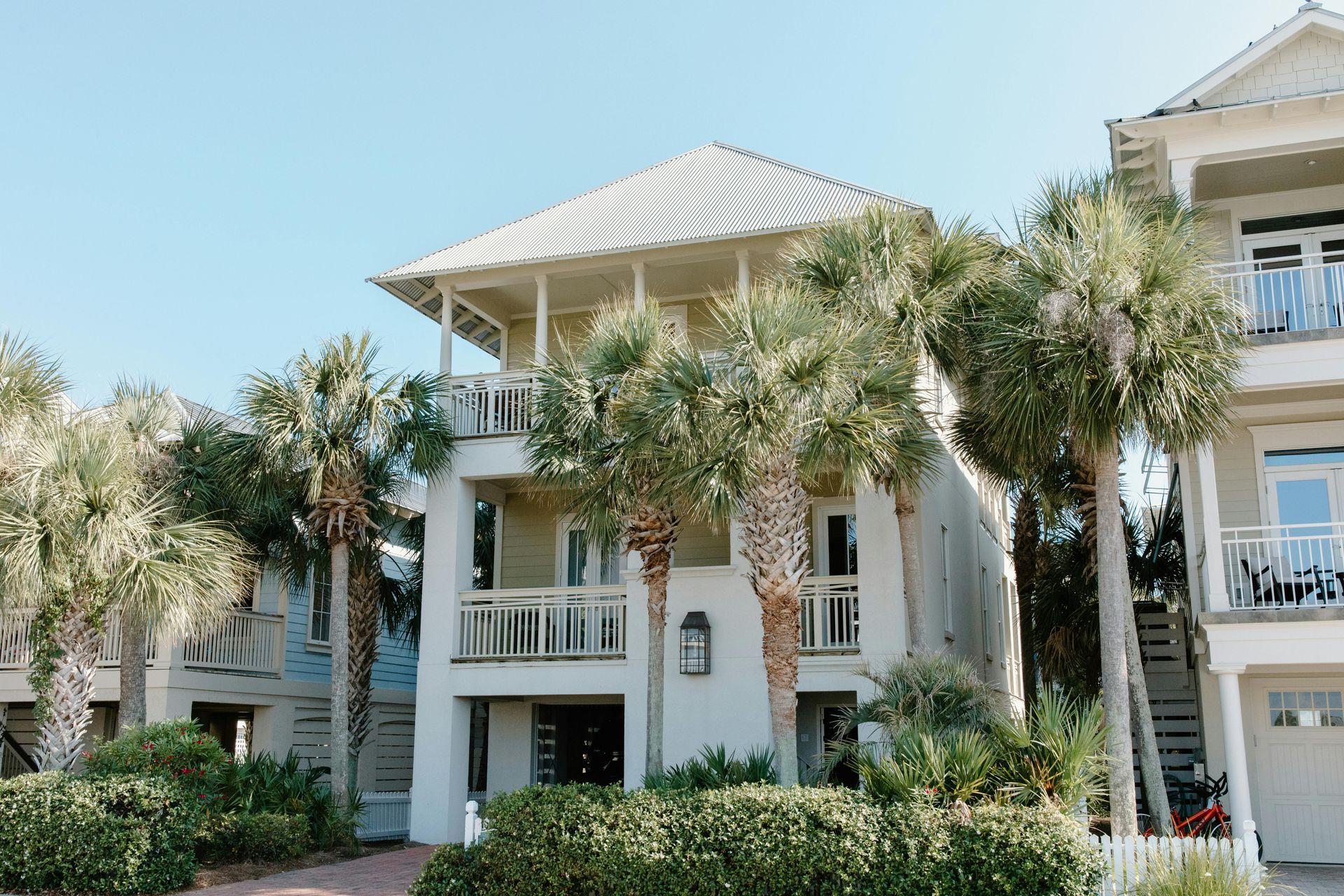 Two-story beach house with balconies, surrounded by palm trees and greenery under a blue sky.